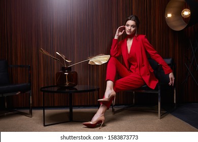 Beautiful Young Pretty Woman Bright Evening Makeup Of Shiny Red Lipstick Brunette Curly Hair Wear Elegant Suit Red Trousers Blouse Jacket High Heels Clothes For Party Meeting Interior Room Fashion.