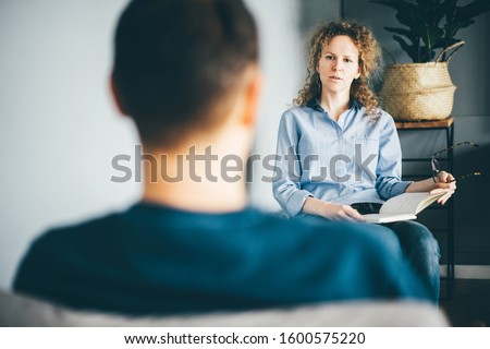 Beautiful young psychotherapist is making notes, and listening man. Psychotherapist consulting man, asking questions, interviewing man sitting on chair. Psychologist with client.