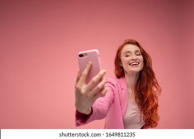 Beautiful young redhead woman making selfie. Funny smiling girl wearing pink jacket holding pink smartphone and posing for selfie isolated on pink background. - Shutterstock ID 1649811235