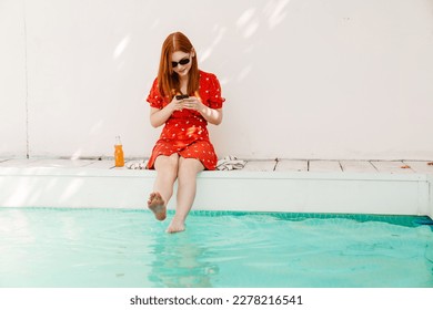 Beautiful young redhead woman wearing red dress using mobile phone while resting by the swimming pool - Powered by Shutterstock