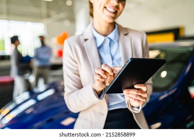 Beautiful Young Saleswoman Working At Car Showroom. Customers In The Background. 