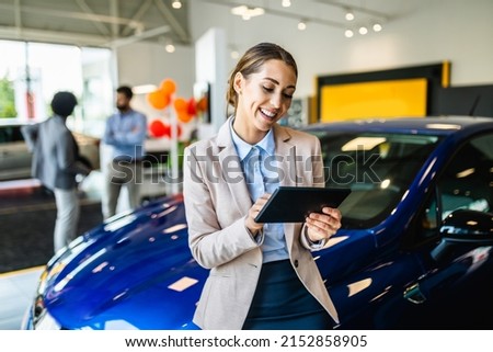 Beautiful young saleswoman working at car showroom. Customers in the background. 