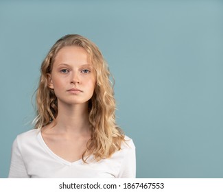 Beautiful young serious clever blonde woman without makeup on color neutral tone Aqua Menthe wall. Pretty female with curly hair in white t shirt