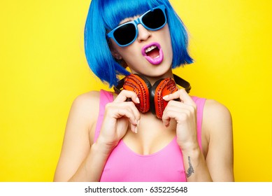 Beautiful young sexy girl dj in blue wig blue sunglasses and pink bathing suit having fun and listening to music in red headphones on yellow background