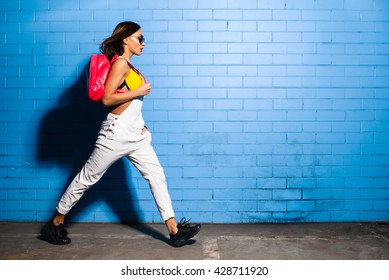 Hermosa joven hipster sexy se acerca al fondo de la pared azul urbana en traje de baño amarillo, traje de salto blanco, zapatillas de deporte, gafas de sol, mochila rosa.