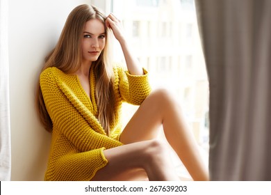 Beautiful young sexy woman with long blonde hair with natural make-up wearing Sweater sun tan body skin pure natural sitting next to window model with clothing catalog spring collection fashion style 
