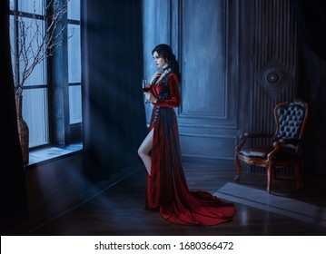 Beautiful young sexy woman vampire in medieval dark castle. Red long gothic dress. Black wavy hair. Backdrop vintage room interior. Hold glass blood wine. image queen of night horror holiday halloween