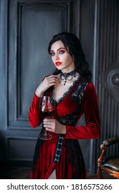Beautiful young sexy woman vampire in medieval dark castle. Red gothic evening dress. Black wavy hair. Backdrop vintage room. A girl holding a glass of bloody wine in her hands. Image queen of night.
