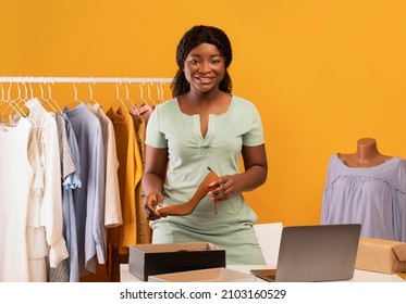 Beautiful Young Shop Assistant Holding Elegant Shoe At Clothes Showroom, Standing Near Table With Laptop Pc, Receiving Online Orders, Selling Outfits Over Internet On Orange Studio Background