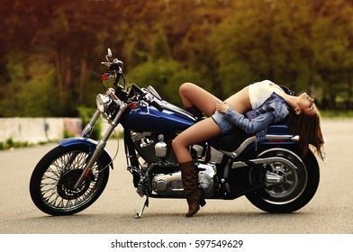 A beautiful young slender girl (woman) with long hair, dressed in a denim jacket, shorts and brown boots lies on a motorcycle. Portrait in profile, full-length. Shooting outdoors