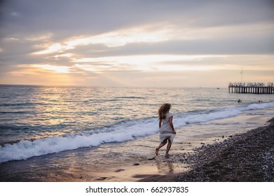 Beautiful young slim girl running on a sandy beach by sea. In a long white dress. Waves. Against the background of the sea. Turkey. Vacation. Sunlight. Place for text. Night. Sunset. horizon. back