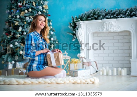Beautiful young slim smiling pregnant woman in a plaid shirt open unpacking gifts near the Christmas tree and fireplace.