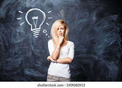 Beautiful Young Smart Blonde Woman Is Thinking Near The Chalk Board With A Drawn Light Bulb. Creative Idea Concept