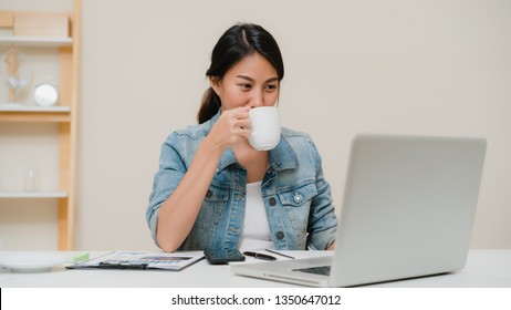 Beautiful young smiling asian woman working on laptop and drinking coffee in living room at home. Asia business woman working document finance and calculator in her home office. Enjoying time at home. - Powered by Shutterstock