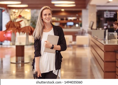 Beautiful Young Smiling Caucasian Woman Hotel Manager Posing In The Lobby. Woman IT Developer With Tablet Stands In The Lobby Of Her Large Company. Successful Beautiful Woman Concept
