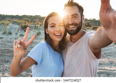 Beautiful young smiling couple spending time at the beach, embracing while taking a selfie, showing peace gesture - Powered by Shutterstock