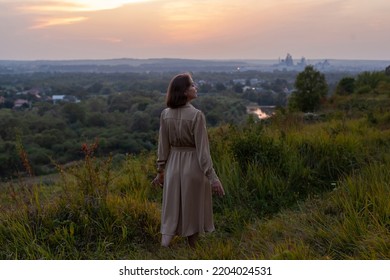 Beautiful young smiling girl in a long brown dress stands along the lawn. Happy woman walks at sunset on a hill overlooking the river. Concept of having rest in park during summer holidays or weekends