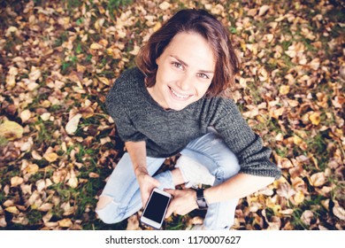 Beautiful young smiling girl woman with wavy hair sitting on grass and on fallen autumn leaves with smartphones in hands, shot from above, portrait with look at camera - Powered by Shutterstock