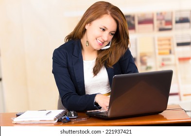 Beautiful Young Smiling Girl Working Behind A Desk With Laptop Talking On Cell Phone