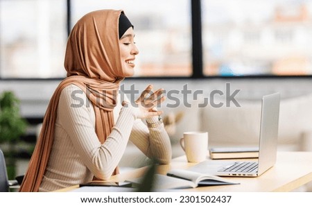beautiful young smiling muslim woman in traditional religious hijab working remotely behind a laptop,  communicates by video call at home
