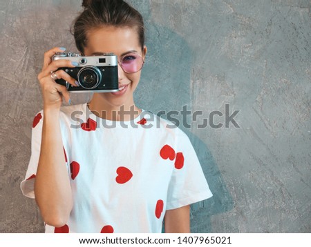 Beautiful young smiling photographer girl taking photos using her retro camera. Woman making pictures. Model dressed in casual summer white t-shirt. Posing In studio near gray wall