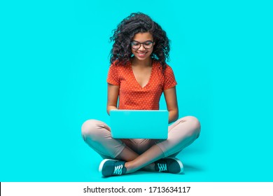 Beautiful Young Smiling Woman In Casual Outfit And Trendy Eyeglasses Sitting Isolated On Bright Colored Blue Background And Working On Her Laptop. Full Length Horizontal Studio Shot. 