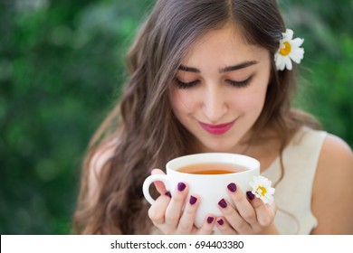 A beautiful young smiling woman with long curly hair and long eyelashes in white top and a flower in hair holding and looking down at the white cup of tea in hands, with green trees in the background - Powered by Shutterstock