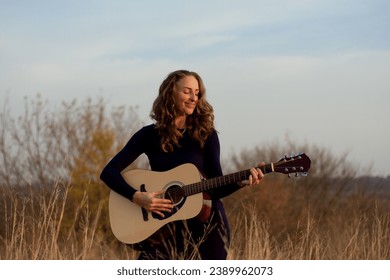 beautiful young smiling woman playing an acoustic guitar while standing in field among tall dried grass. Talented musician. Autumn sunset. Romantic mood. Creativity, lifestyle - Powered by Shutterstock