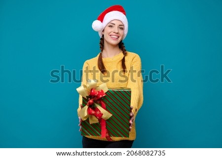 Beautiful young smiling woman in santa claus hat holding a new year gift on a blue isolated background. New year concept