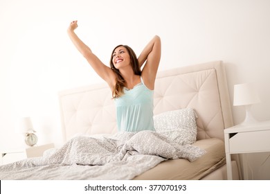 Beautiful Young Smiling Woman Sitting On Bed And Stretching In The Morning At Bedroom.