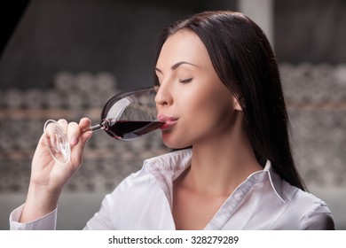 Beautiful Young Sommelier Is Drinking Red Wine In Cellar. The Woman Is Standing And Holding Wineglass. She Closed Eyes With Pleasure