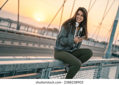 Beautiful young sportswoman smiling while holding a phone outdoors. Young woman sitting after workout during the sunrise in urban environment. - Powered by Shutterstock