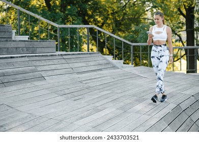 Beautiful young sporty woman walking with smartphone on the wooden pier or stairs in the park. Smilling fitness girl smiling and looking to the phone
