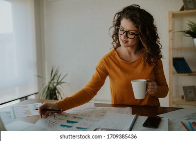 Beautiful young statistician is analyzing data while having coffee at office. Young business woman is enjoying her work.