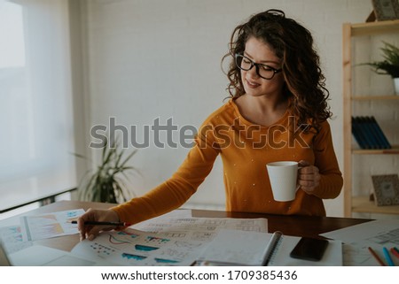 Beautiful young statistician is analyzing data while having coffee at office. Young business woman is enjoying her work.