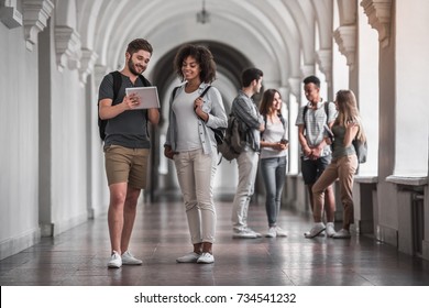 Beautiful Young Students Are Resting In University Hall, A Couple In The Foreground Is Using A Digital Tablet And Smiling