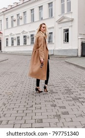 Beautiful Young Stylish Blonde Woman Wearing Long Beige Coat And Black High Heel Shoes Walking Through The City Streets. Trendy Casual Outfit. Street Fashion. Back View.