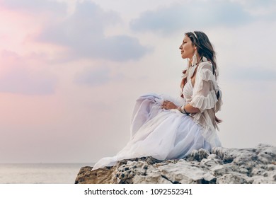 beautiful young stylish boho woman sitting on the beach at sunset