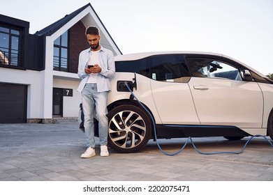 Beautiful young stylish man is with electric car at daytime charging the vehicle and holding smartphone. - Powered by Shutterstock