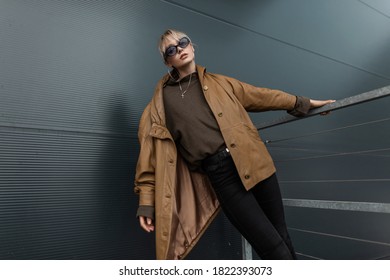Beautiful young stylish woman with fashionable sunglasses in a retro brown leather jacket with a sweater and black jeans posing on the street
