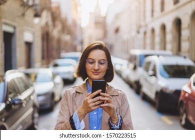 Beautiful young stylish woman walking in the old european city streets using modern smartphone