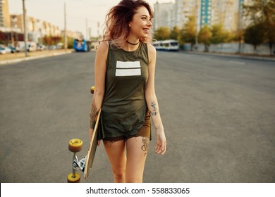 Schöne junge tätowierte Frau mit seinem Langboard auf der Straße in der Stadt bei sonnigem Wetter