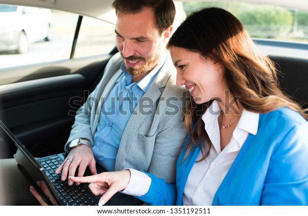 Beautiful young team of two business\
people driving on a backseat of a car and working on laptop and\
smiling. Successful team businessman and businesswoman.\
