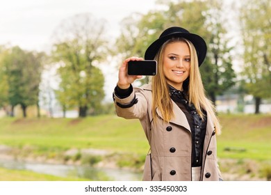 Beautiful Young Teenage Girl With Long Blonde Hair In Fedora And Trench Coat Taking A Selfie On A Smartphone In Autumn In Park. Horizontal, Mild Retouch, Vibrant Colors.