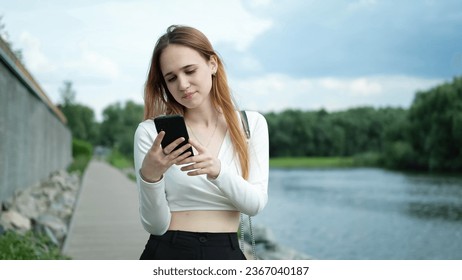 A beautiful young teenage girl in a white top, with long brown hair, uses a smartphone outdoors in the summer, while walking in nature on the shore of a lake. The girl writes text messages and - Shutterstock ID 2367040187