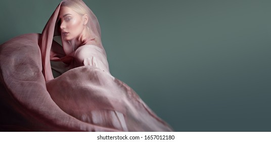 A beautiful young tender girl in a chiffon dress is covered with a drapery made of thin fabric that develops in the wind.