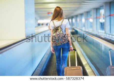Beautiful young tourist girl with backpack and carry on luggage in international airport, on travelator