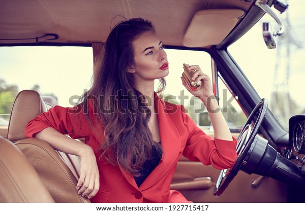 Beautiful young vogue woman in red suit sitting\
in retro car using a\
perfume.