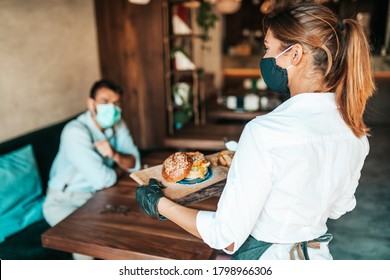 Beautiful Young Waitress With Face Protective Mask Serving Delicious Burger To Middle Age Male Customer. Corona Virus And Small Business Is Open For Work Concept.