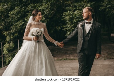 Beautiful Young Wedding Couple In A Green Summer Park. Brides At A Photo Shoot. The Bride In A White Modern Dress And Veil, A Man In A Classic Grey Suit With A Bow Tie. Happy Wedding Day, Love, Hugs.